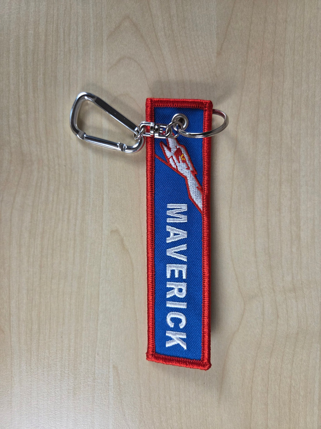 Keychain with 'MAVERICK' text on a blue background, attached to a metal carabiner, on a wooden surface.