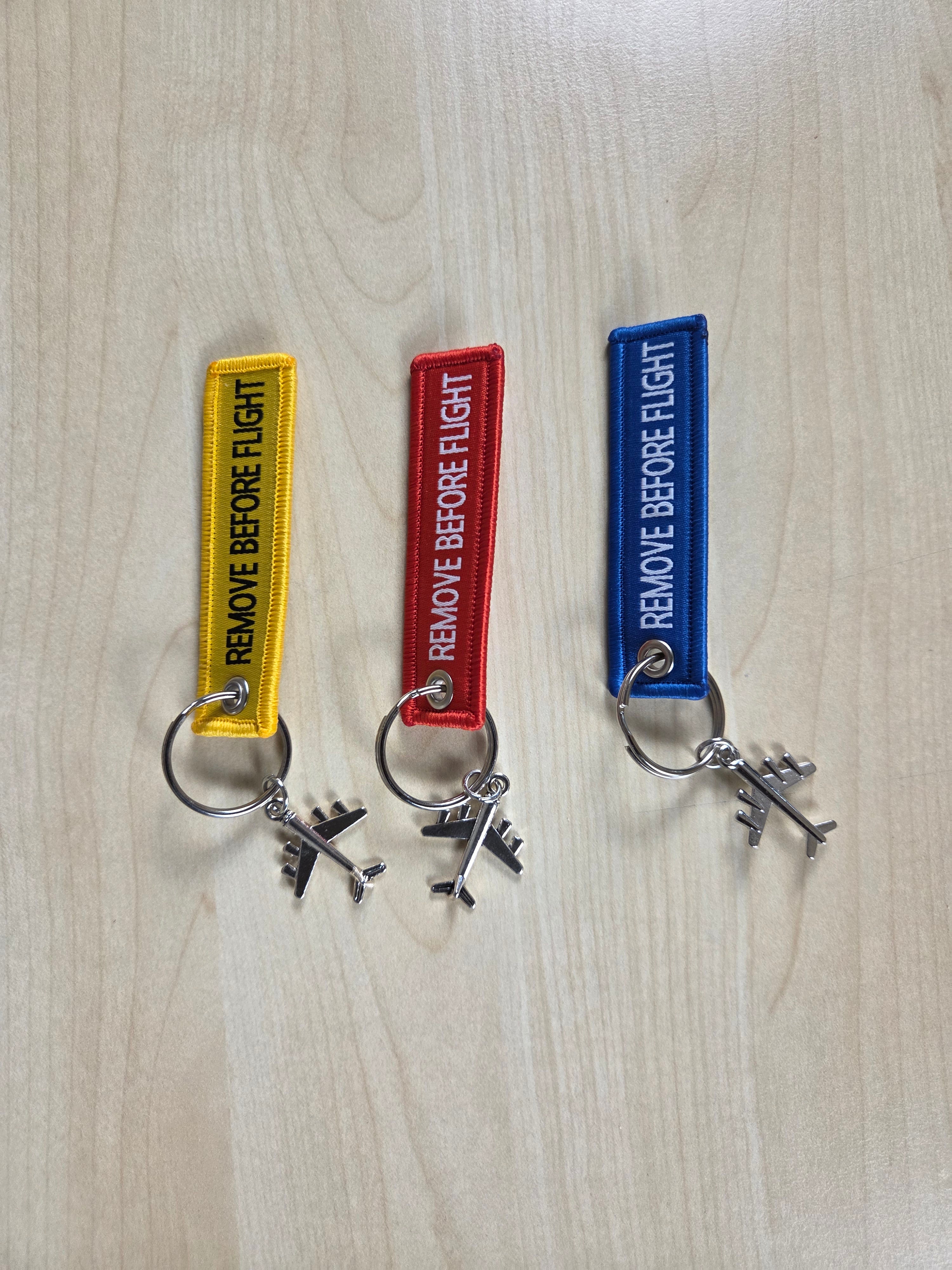 Three embroidered keychains with silver airplane charms are displayed on a light wood grain surface. The keychains are yellow, red, and blue, each featuring a vertical rectangular patch with the text 'REMOVE BEFORE FLIGHT' in white capital letters.