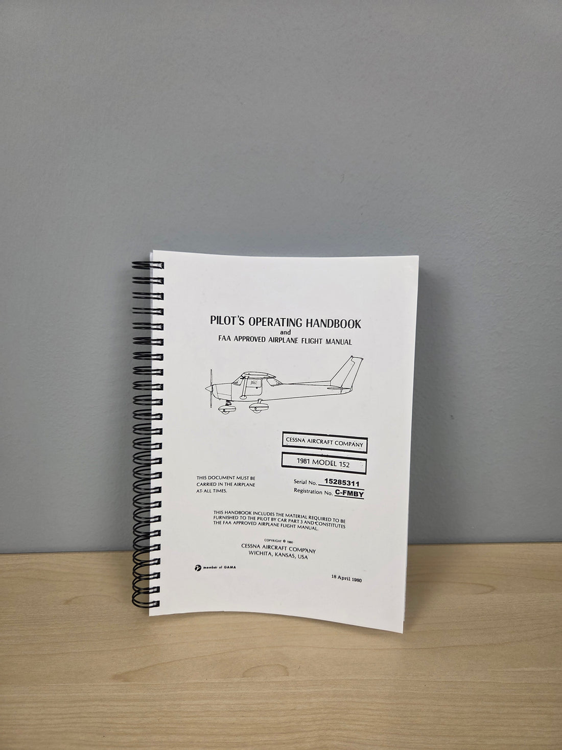 A spiral-bound Pilot's Operating Handbook for a Cessna 172 aircraft, displayed standing upright on a light wood surface against a neutral gray wall.