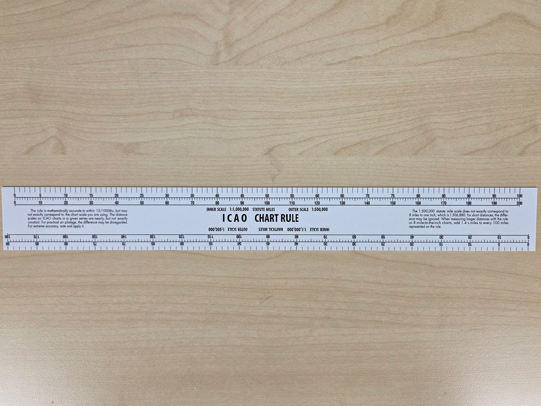 A white plastic 12 x 6 inch aviation chart ruler with printed measurement markings, shown vertically on a light wood grain surface.