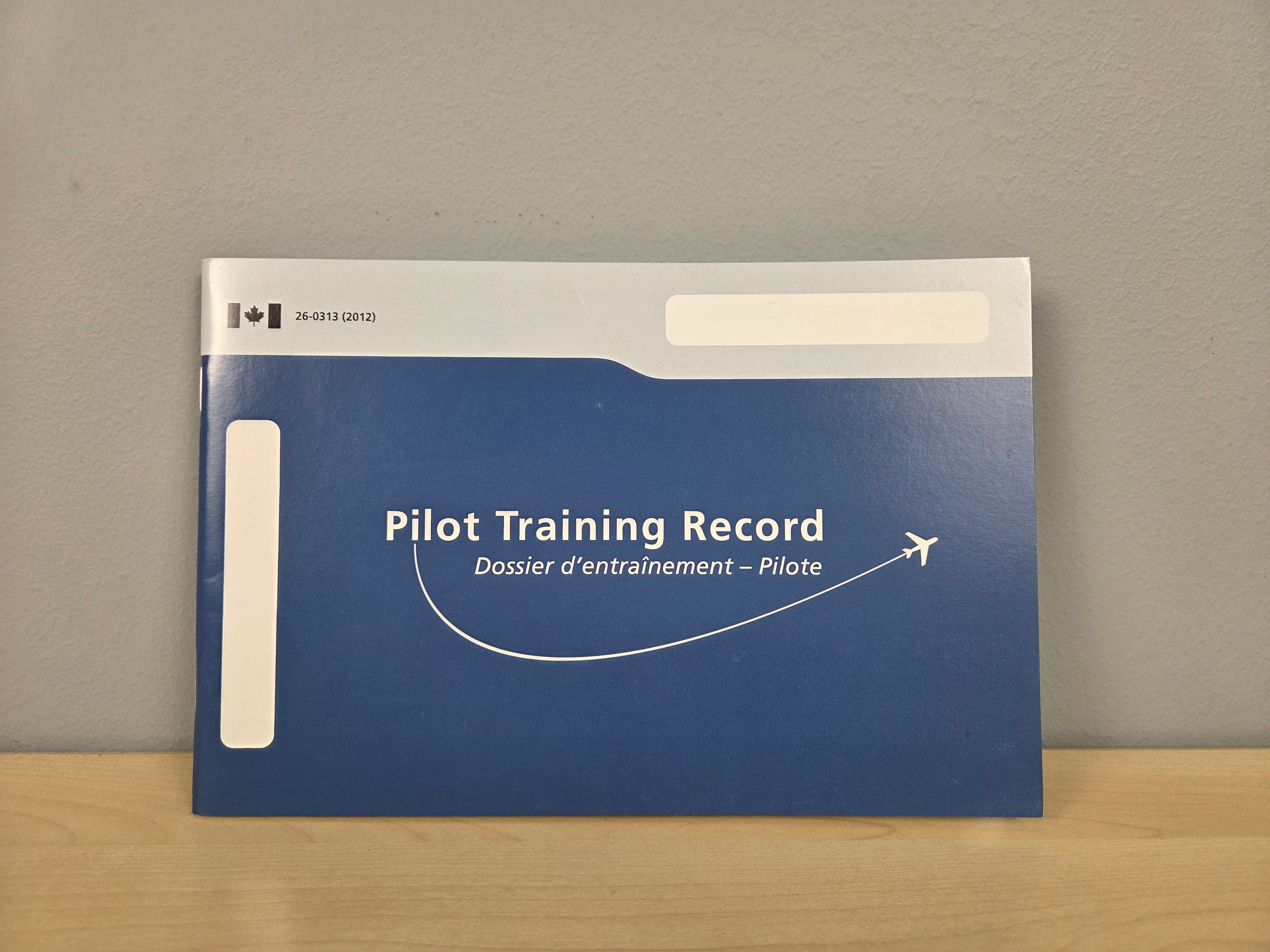 A Pilot Training Record folder with a blue cover and white text, standing on a light wood surface against a gray wall.