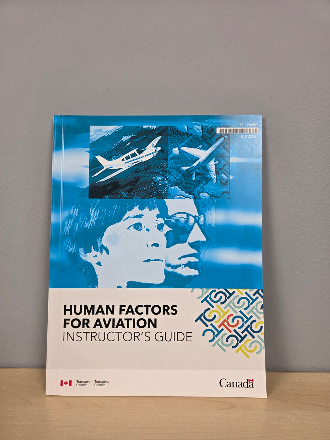 A book titled 'Human Factors for Aviation Instructor's Guide' standing upright on a light wood surface against a gray wall. The cover features a blue-toned graphic with two pilots' profiles and an airplane photo, plus a multicolor patterned section on the lower right.