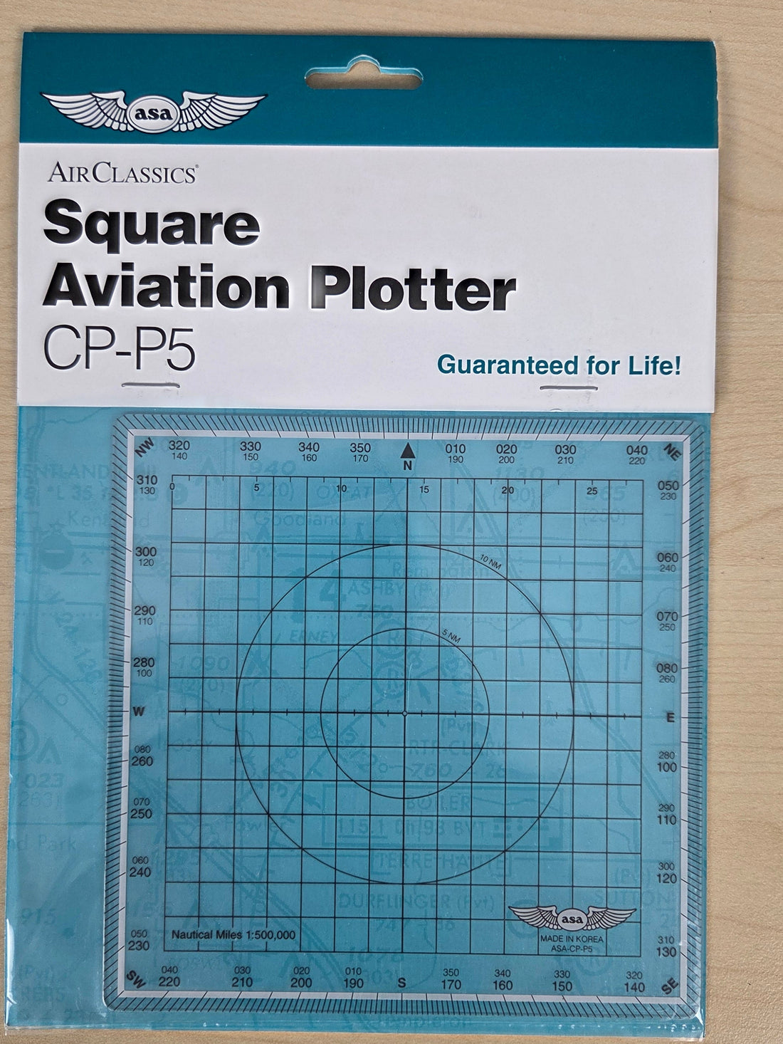 AirClassics Square Aviation Plotter CP-P5 in its original retail packaging, displayed on a light wood surface. The product is a square grid plotter with concentric circles, shown through a clear window in a teal and white cardboard backing.