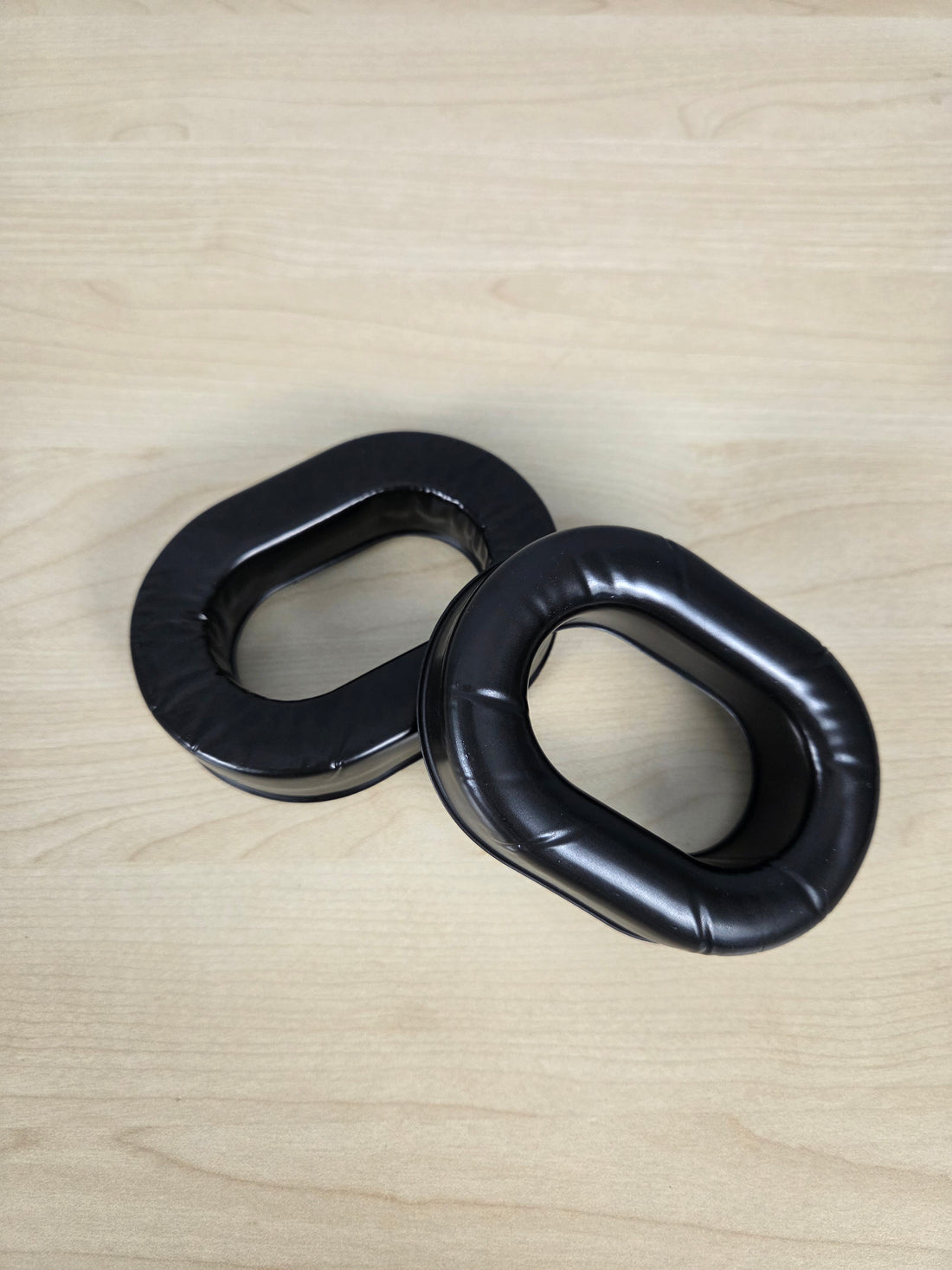 Two black padded looped foam or rubber pieces, replacement ear cushions for aviation headset components, placed on a light wood surface.