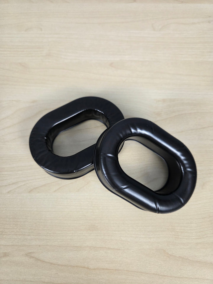 Two black padded looped foam or rubber pieces, replacement ear cushions for aviation headset components, placed on a light wood surface.