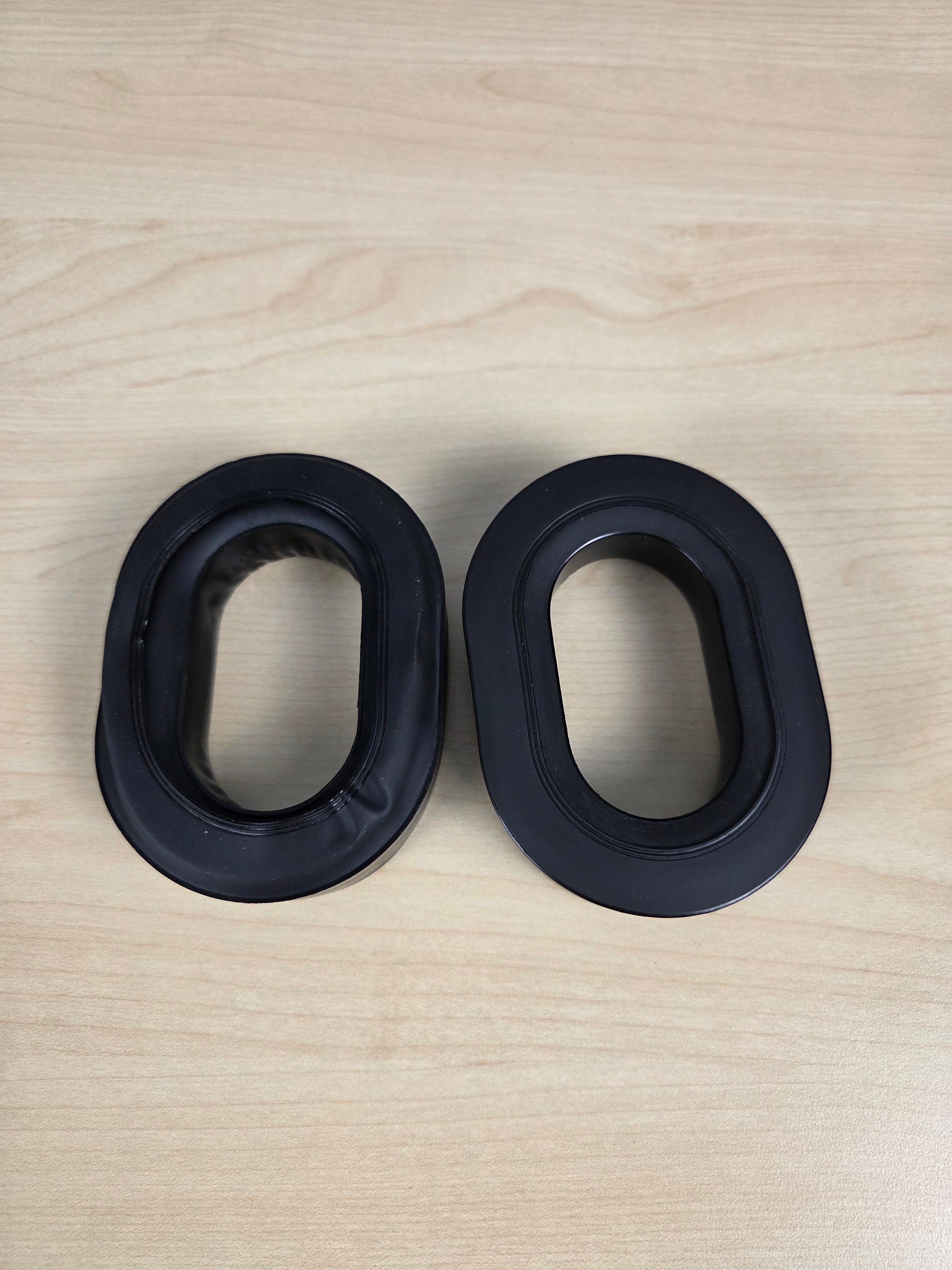 Two black oval-shaped silicone or rubber aviation headset pads placed side by side on a light wood grain surface.