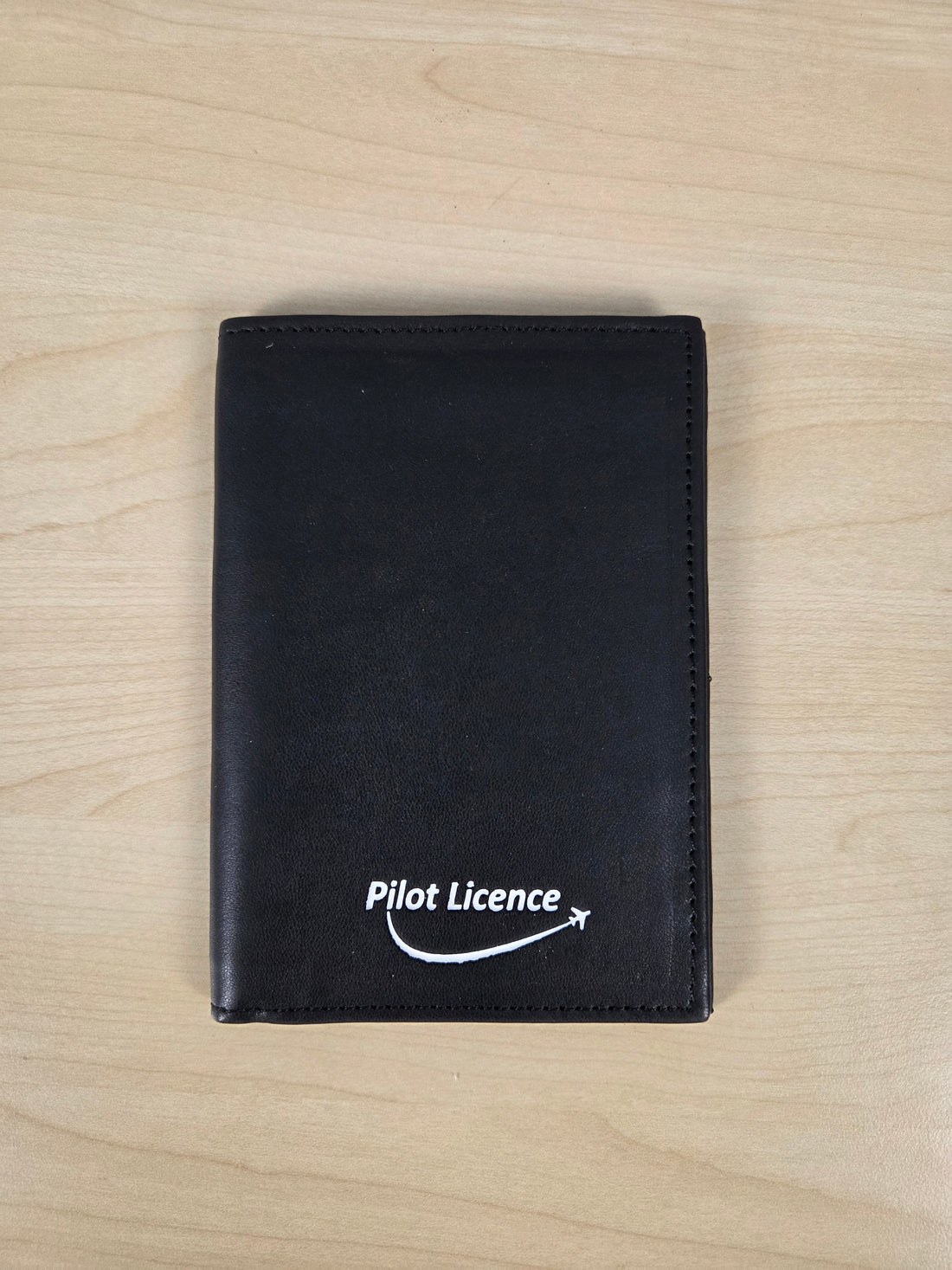 A black leather wallet with a white 'Pilot Licence' logo printed on the front, displayed flat on a light wood grain surface.
