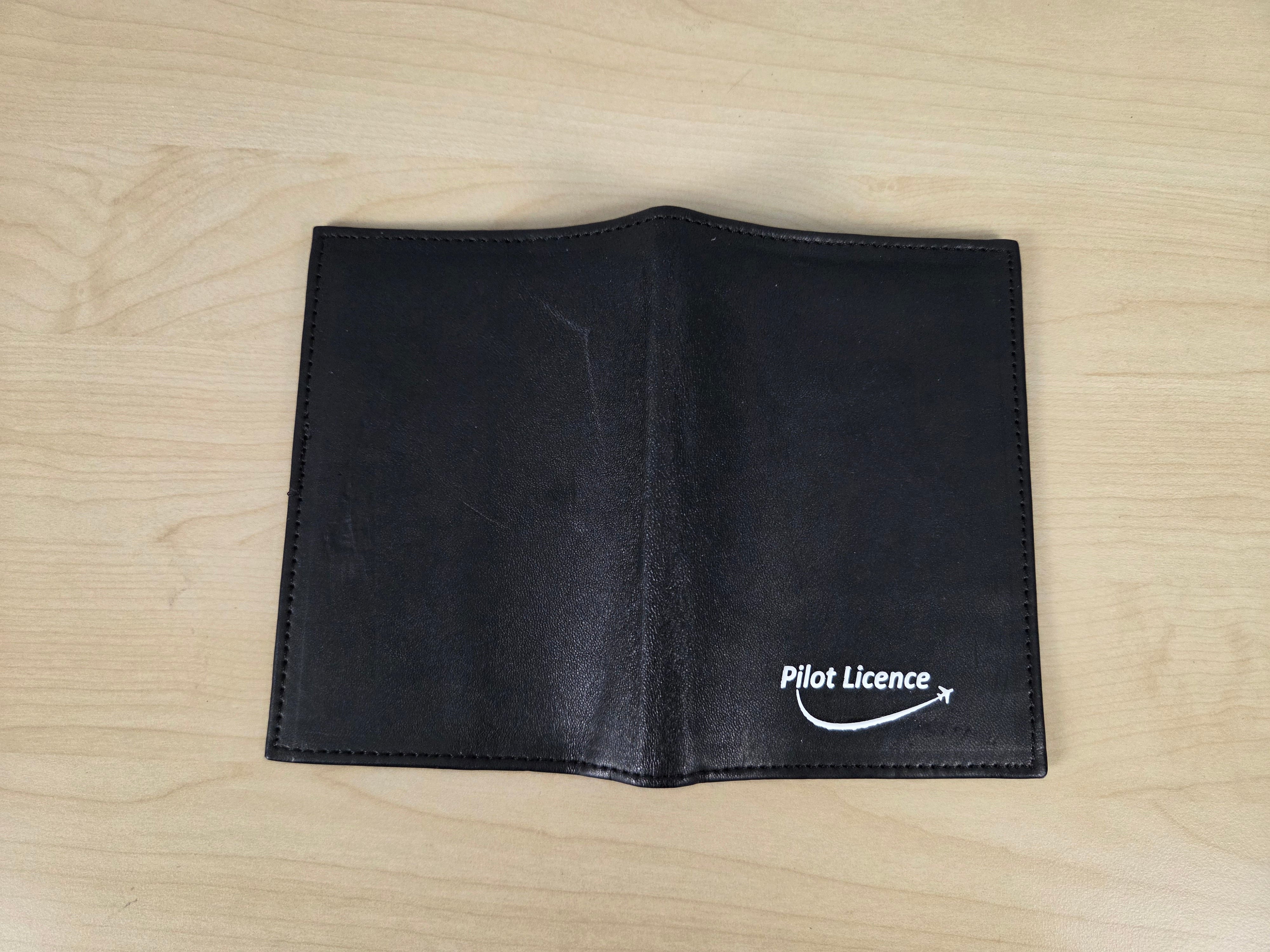 A black leather bifold wallet with 'Pilot Licence' printed in white on the left panel, displayed flat on a light wood grain surface.