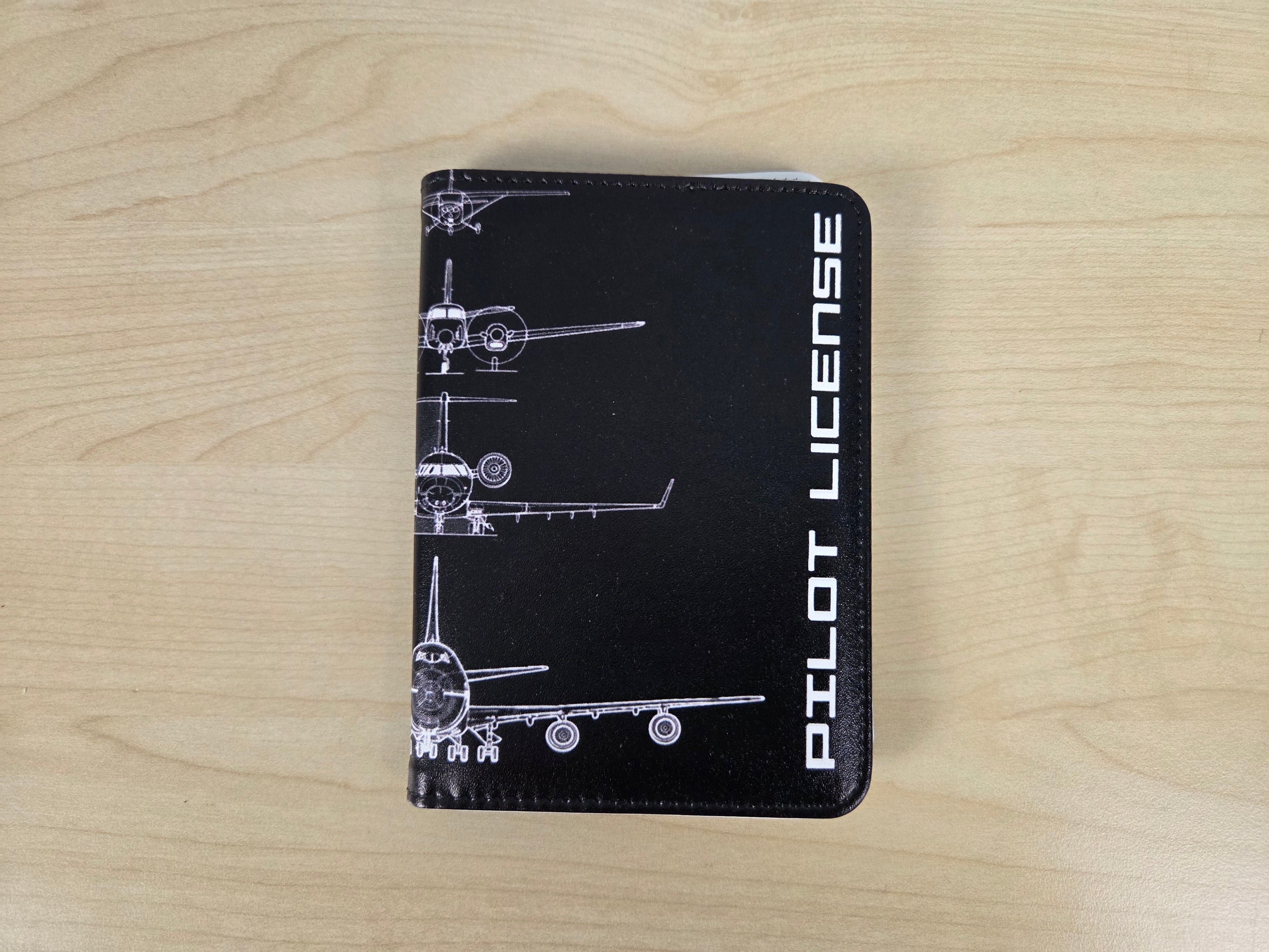 A black leather passport cover featuring white airplane schematics and the vertical text 'PILOT LICENSE' on the left side, displayed on a light wood grain surface.