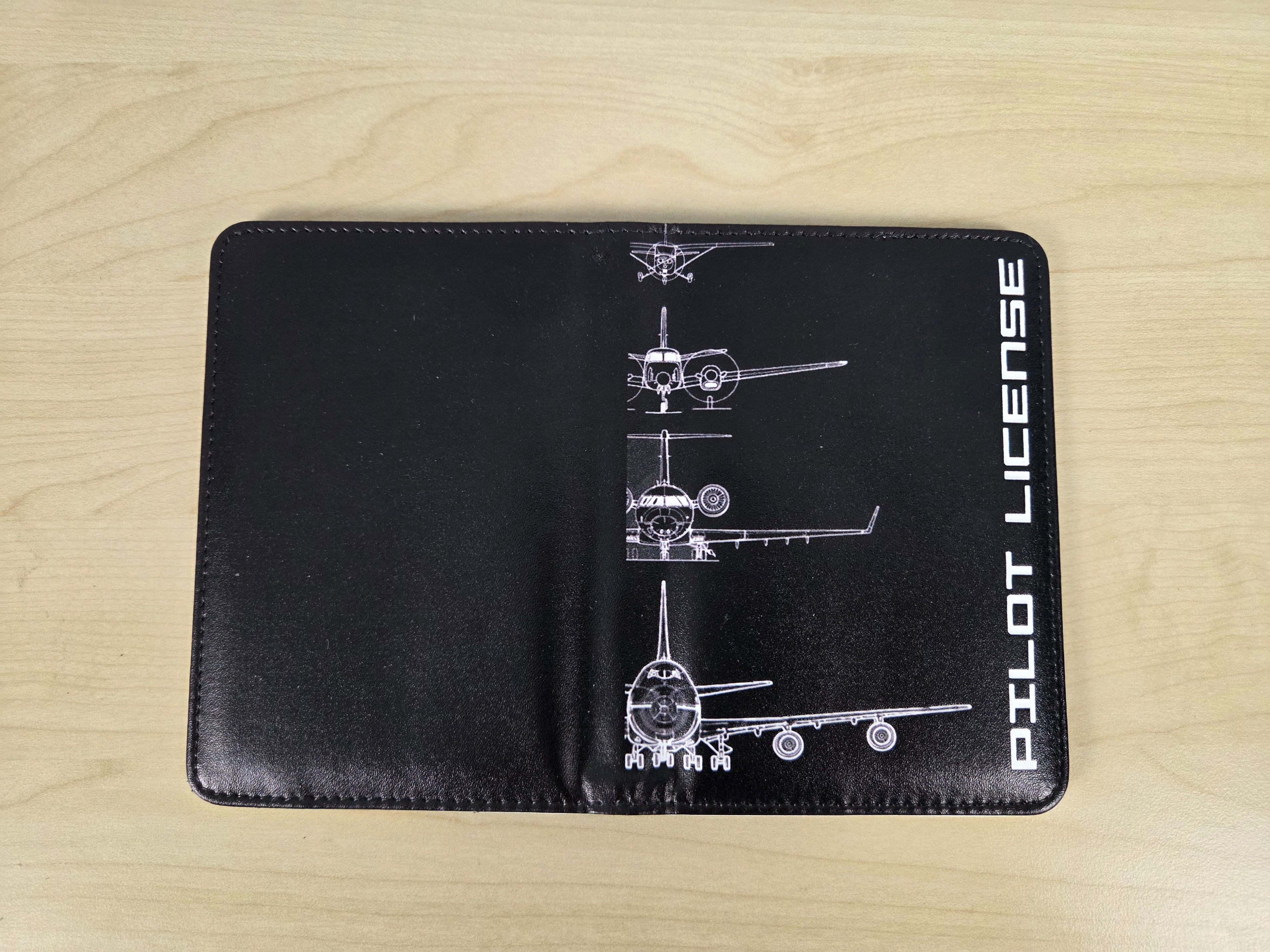 A black leather passport cover featuring white airplane schematics and the vertical text 'PILOT LICENSE' on the left side, displayed flat on a light wood background.