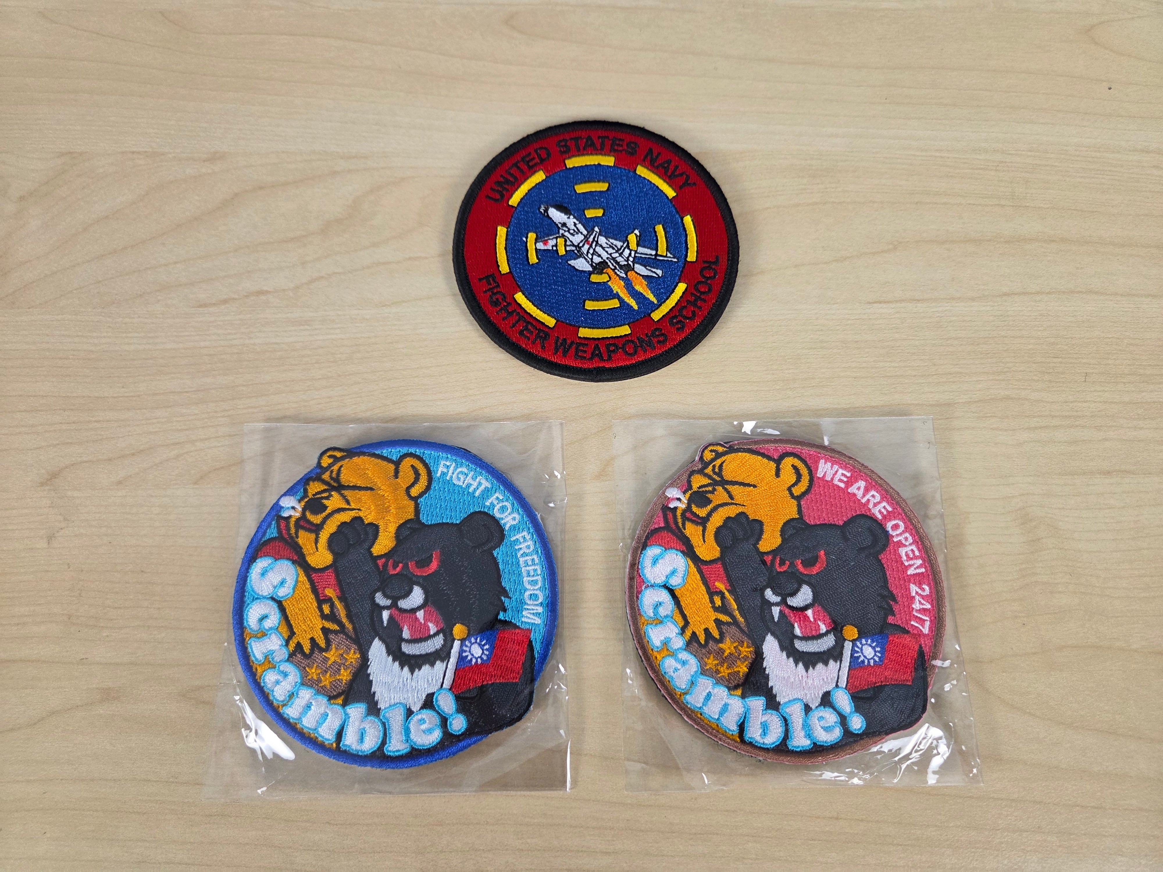 Three embroidered military-style patches displayed on a light wood surface: two patches still in clear plastic packaging and one patch laid flat. The patches feature a black bear, yellow tiger, red and blue background elements, and visible text including 'Scrubdole' and '306th Support Battalion'.