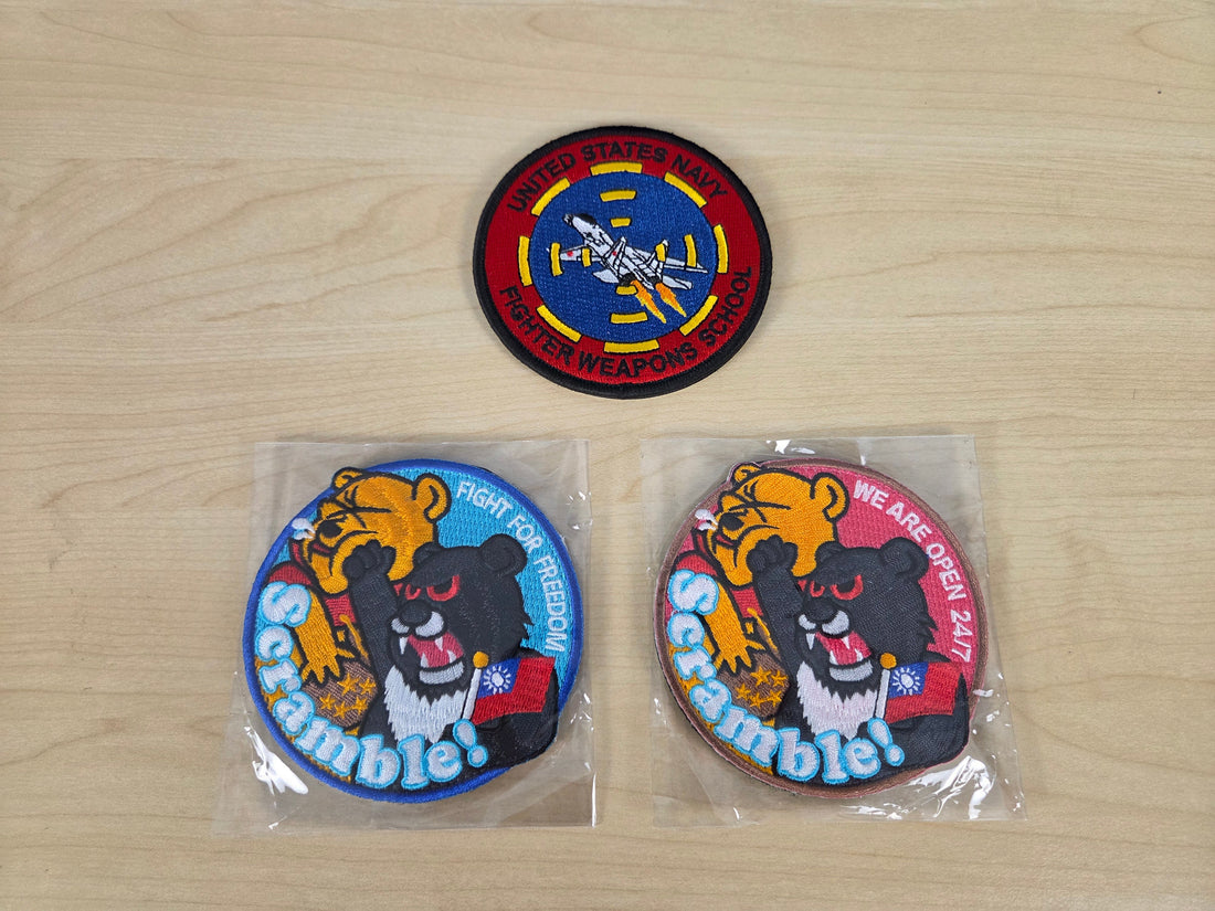 Three embroidered military-style patches displayed on a light wood surface: two patches still in clear plastic packaging and one patch laid flat. The patches feature a black bear, yellow tiger, red and blue background elements, and visible text including 'Scrubdole' and '306th Support Battalion'.