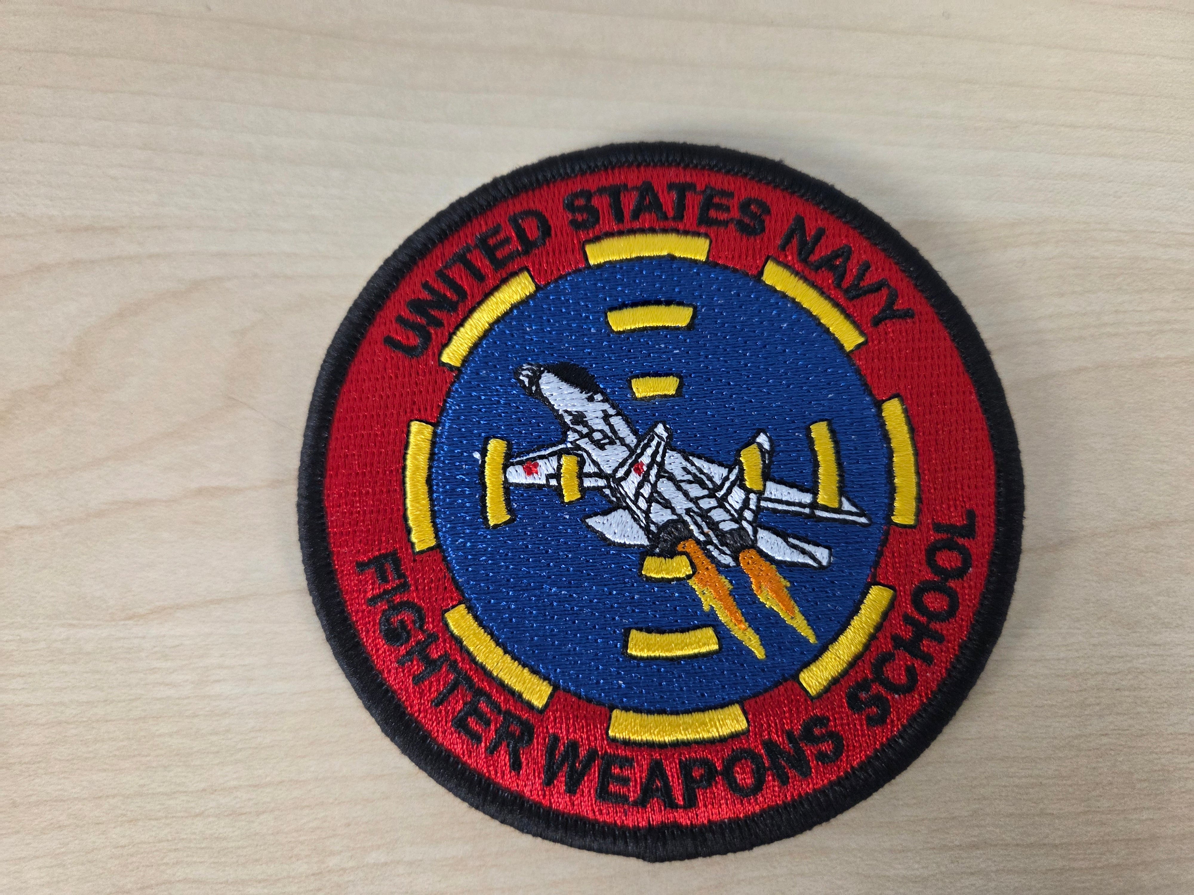 An embroidered circular patch for the United States Navy Fighter Weapons School, featuring a white fighter jet on a blue background with a red outer ring and yellow accents, displayed on a light wood surface.
