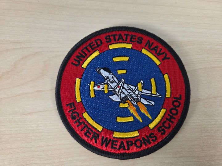 An embroidered circular patch for the United States Navy Fighter Weapons School, featuring a white fighter jet on a blue background with a red outer ring and yellow accents, displayed on a light wood surface.