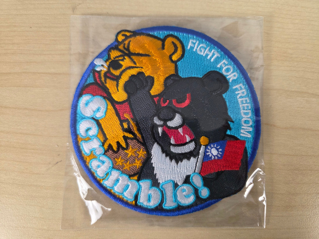 A round embroidered patch in clear plastic packaging, featuring cartoon skunks and a tiger with the text 'Scramble! Fight for Freedom' on a light wood background.