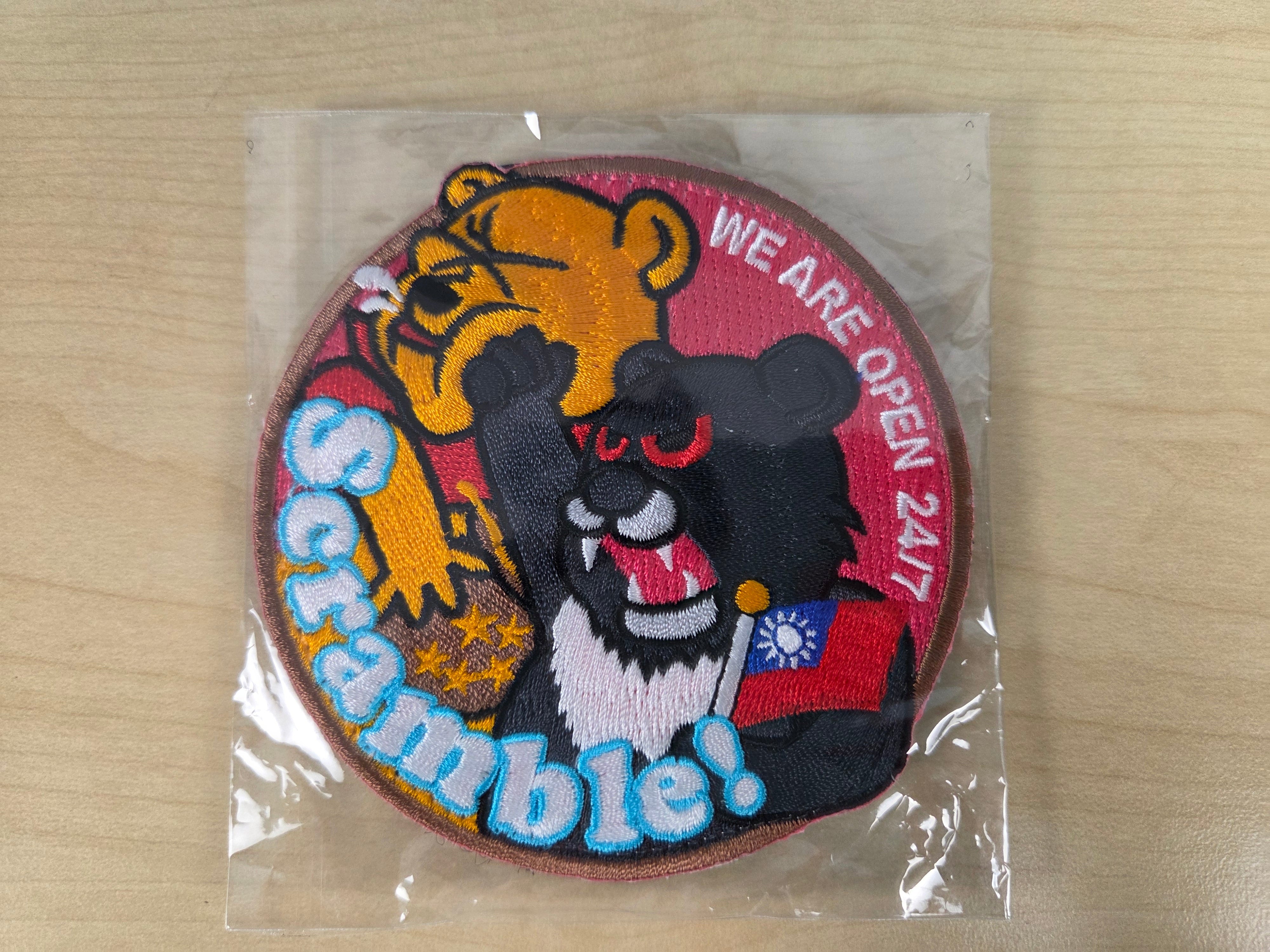 A round embroidered patch in clear plastic packaging, featuring cartoon characters and the text 'Scramble!' and 'WE ARE OPEN 24/7' on a light wood surface.