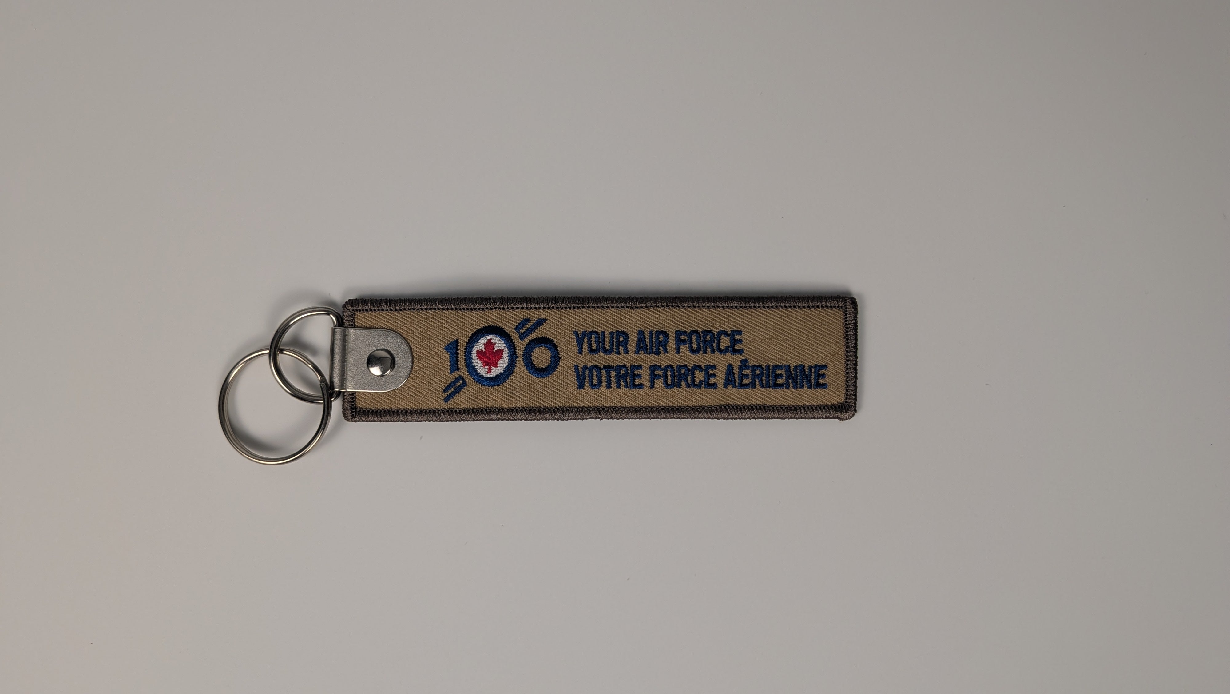 A rectangular tan leather keychain with a silver split ring, featuring the text 'YOUR AIR FORCE VOTRE FORCE AÉRIENNE' and a '100' with a red maple leaf emblem, on a neutral gray background.