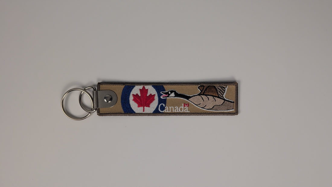 A rectangular woven Canada keychain strap with a metal split ring, featuring a red maple leaf, a white maple tree, and the word 'Canada' on a light gray background.