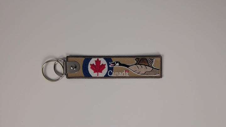 A rectangular woven Canada keychain strap with a metal split ring, featuring a red maple leaf, a white maple tree, and the word 'Canada' on a light gray background.