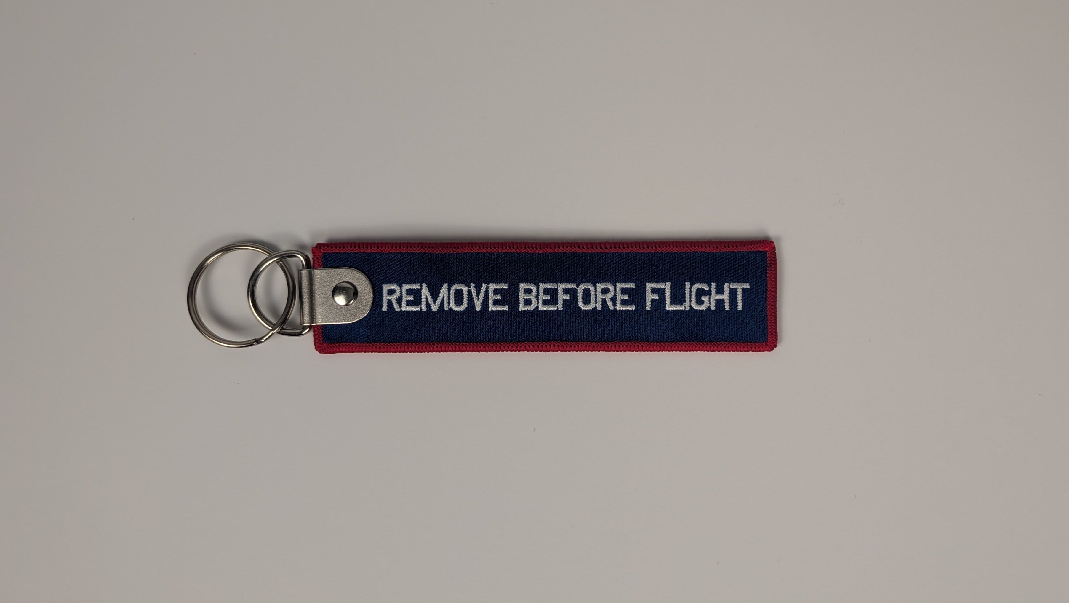 A rectangular navy blue and red fabric keychain with white text reading 'REMOVE BEFORE FLIGHT' attached to a silver metal key ring, shown on a plain light gray background.