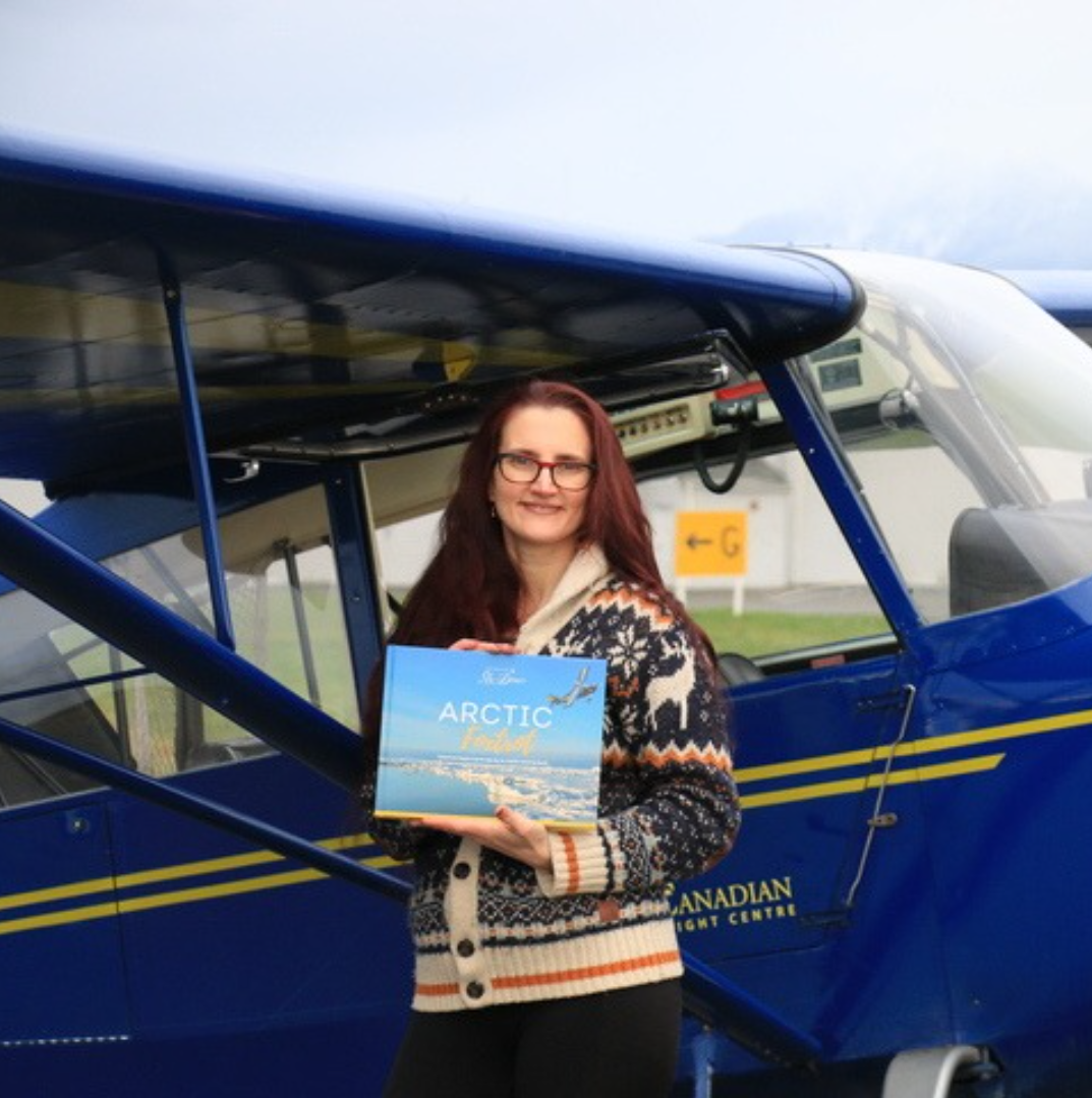 Anna Serbinenko holding a book titled 'Arctic Foxtrot' in front of a blue and white airplane.