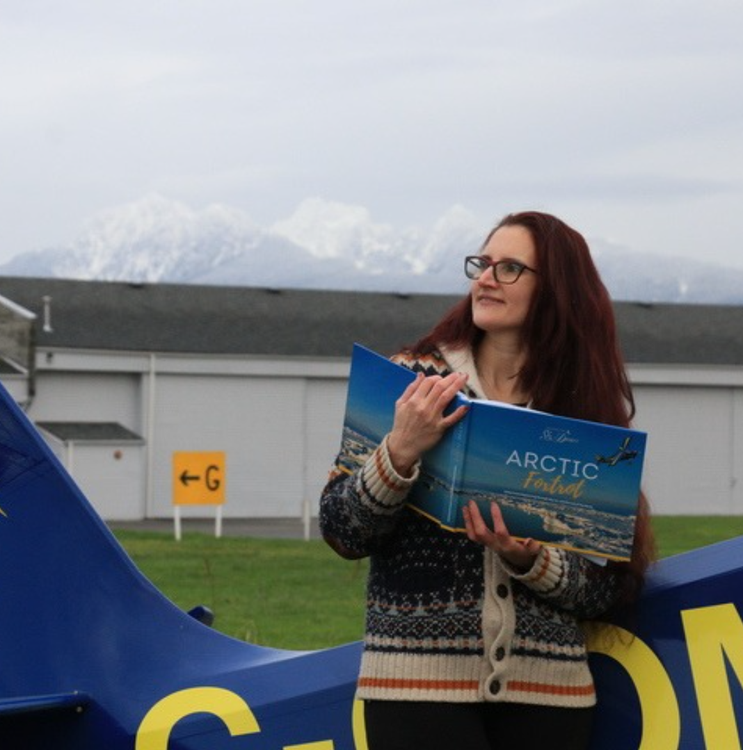 Book author, Anna Serbinenko, holding a book titled 'Arctic' in front of an airplane with mountains in the background